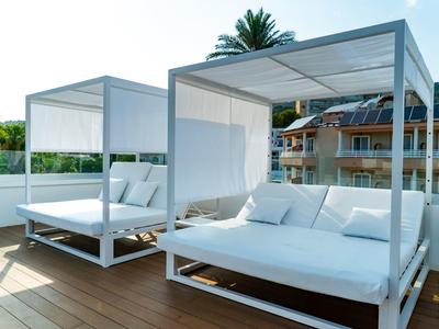 Zwei weiße Outdoor-Sofas mit Überdachung auf Holzterrasse, im Hintergrund grüne Pflanzen und Gebäude.