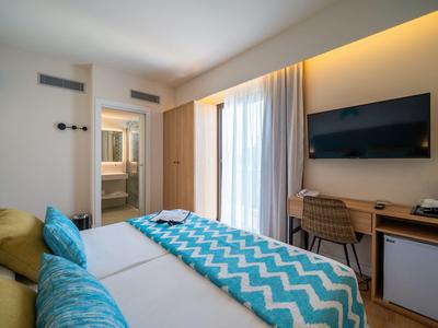 Hell eingerichtetes Schlafzimmer mit Doppelbett, blauen Kissen, Fernseher und Zugang zu Bad.