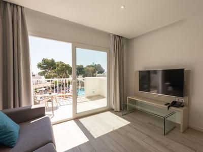 Lichte kamer met bank, tv, glazen tafel en uitzicht op een balkon met zwembad.