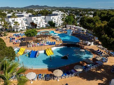 Hotel pool with sun umbrellas and loungers, surrounded by white buildings and green landscape.