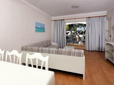 Bright hotel room with sofa, dining table, and balcony overlooking palm trees and pool.