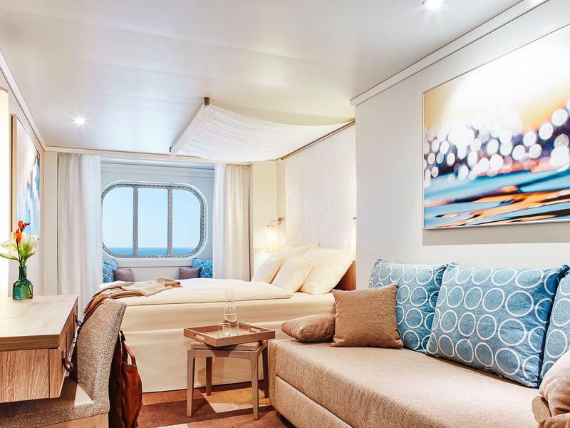 Bright, modern hotel room with large bed, sofa, and porthole window with ocean view.