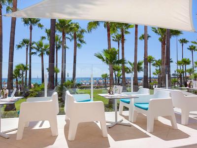 Chaises blanches modernes et table sous une voile d'ombrage avec palmiers et vue sur la mer.