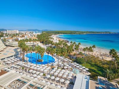 Resort with pool, umbrellas, palm trees, and sandy beach under clear blue sky.