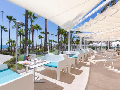 Sunny terrace with white tables and chairs, overlooking palm trees and the sea.