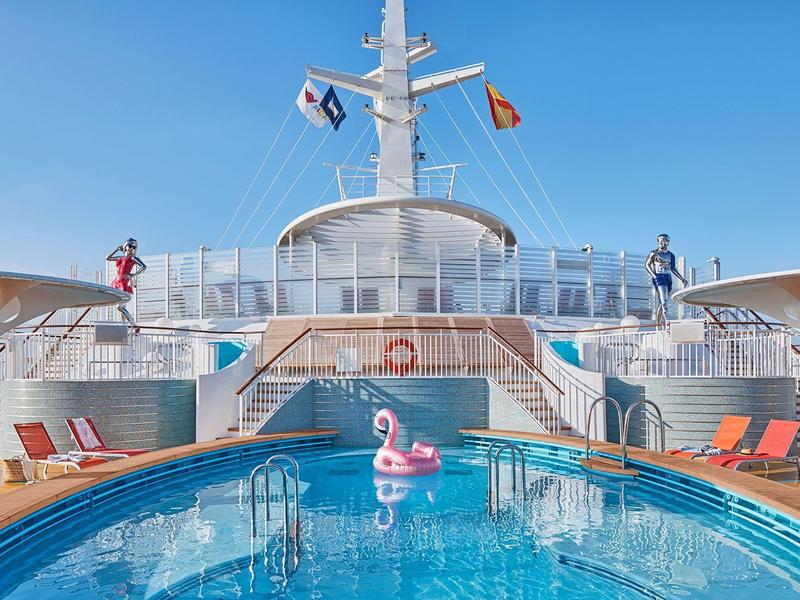 Cruise ship deck with a clear blue pool, pink flamingo float, and lounge chairs under sunny sky.