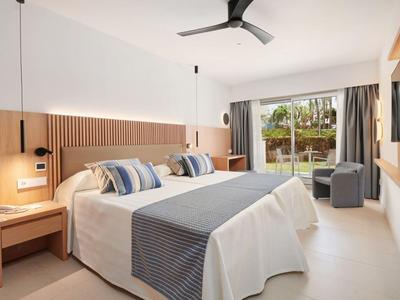 Lichte hotelkamer met tweepersoonsbed, blauwgestreepte kussens, houten decor en schuifpui naar tuin.