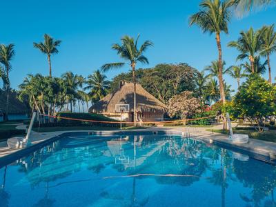 Clear swimming pool with palm trees and a thatched building in the background under a blue sky.