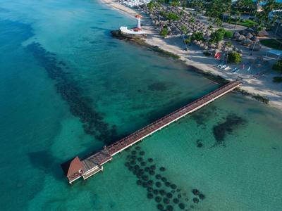 Long pier extends over turquoise water to a coast with a lighthouse and vegetation.