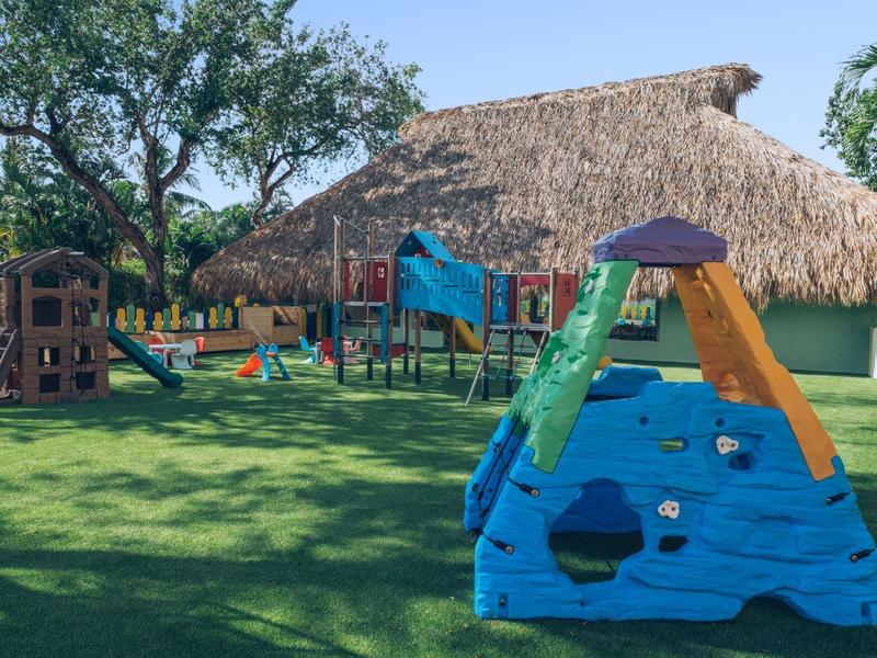 Playground with colorful climbing structures on green grass in front of a thatched building