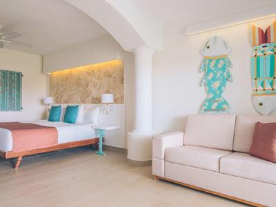 Modern hotel room with double bed, couch, and colorful wall decoration in light tones.
