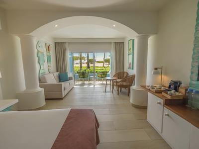Bright hotel room with bed, seating area, and view of terrace with pool and palm trees.