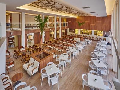 Large modern dining area with white tables, wooden chairs, and palm trees as decoration.