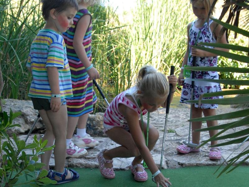 Čtyři děti hrají minigolf v zeleném, slunečném prostředí.