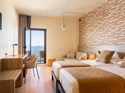 Modern hotel room with two beds, chairs, and a view of the sea through the door