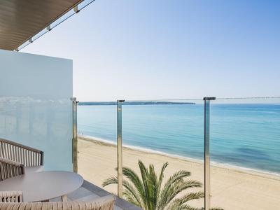 Balcony with wicker chairs and glass railing overlooking a sandy beach and blue sea.