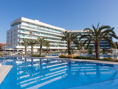 Modern hotel with large pool and palm trees under clear blue sky.