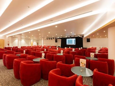 Konferenzraum mit roten Sesseln und Tischen vor einer Bühne mit Bildschirm.