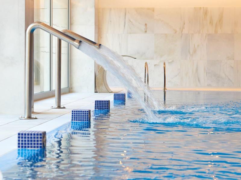 Moderner Innenpool mit Wasserstrahl, blauen Fliesen und Fensterfront.