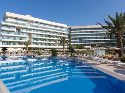 Moderner Hotelpool mit Liegestühlen und Palmen vor einem großen Hotelgebäude bei klarem Himmel.
