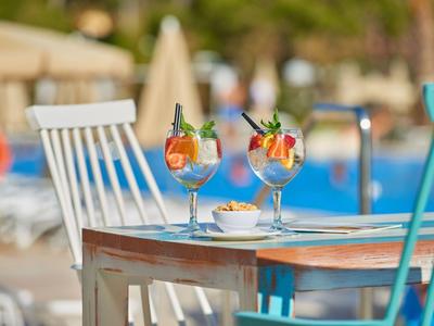 Zwei bunte Cocktails auf einem Holztisch neben einem Pool mit weißen und blauen Stühlen.