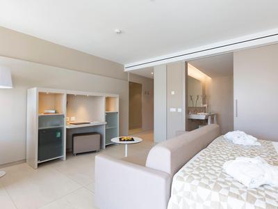 Bright hotel room with double bed, sofa, minibar, and workspace.