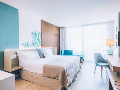 Světý moderní hotelový pokoj s dvojlůžkem, dřevěnou podlahou, psacím stolem a velkým oknem s průsvitnými závěsy.