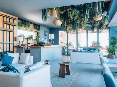 Modern hotel lobby with white sofas, hanging plants, wooden stools, and large windows.
