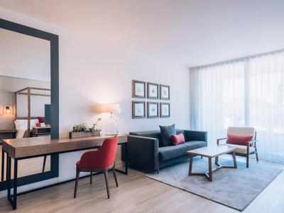 Modern hotel room with sofa, red chairs, large mirror, and natural light through sheer curtains.