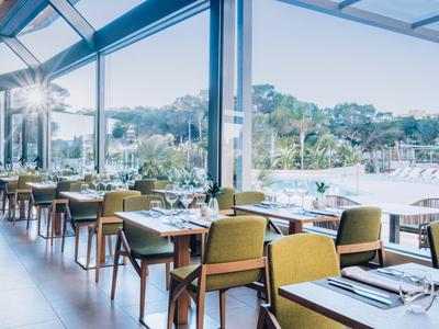 Bright hotel restaurant with large glass windows and green chairs overlooking nature.