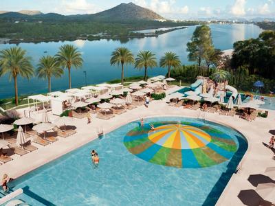 Hotel pool with colorful sun umbrella, surrounded by loungers, palm trees, and mountain view