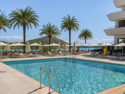 Freizeitpool mit Liegestühlen und Sonnenschirmen neben einem modernen Hotel unter blauem Himmel mit Palmen.