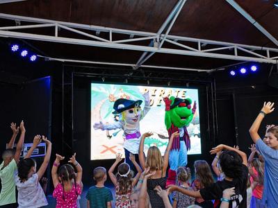 Children stand in front of a stage with two costumed puppets outdoors at an event.