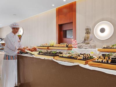 Ein Koch serviert Sushi und andere Speisen an einem Buffet mit einer Buddha-Statue im Hintergrund.