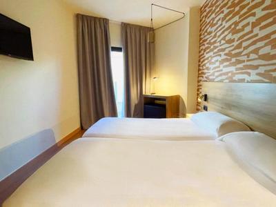 Lichte hotelkamer met twee eenpersoonsbedden, een aan de muur gemonteerde tv en een raam met gordijnen.