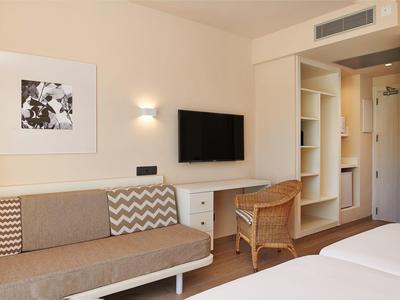 Gemütliches Hotelzimmer mit Sofa, TV, Stuhl und offener Garderobe in neutralen Tönen.