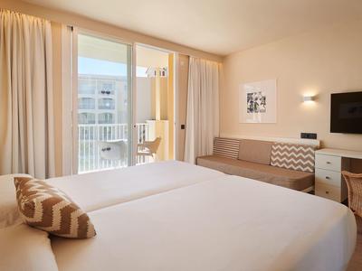 Helles Hotelzimmer mit zwei Betten, Sofa, Flachbildfernseher und Balkon mit Vorhängen.