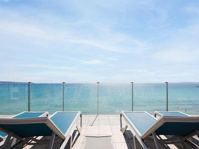 Zwei Liegestühle auf einer Terrasse mit Glasgeländer und Blick auf das blaue Meer unter blauem Himmel.