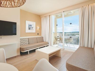 Modern hotelkamer met twee banken, tv, balkon en lichte houten vloer.