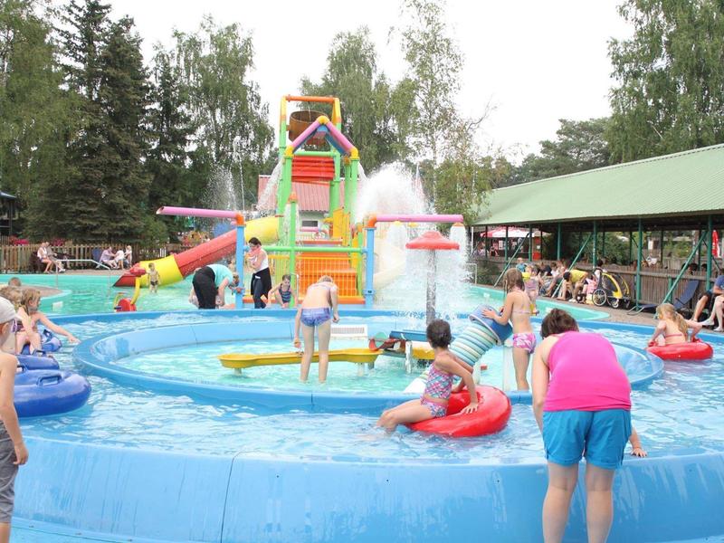 Kinder und Erwachsene spielen in einem runden Planschbecken mit Wasserspielplatz und Rutsche im Freien.