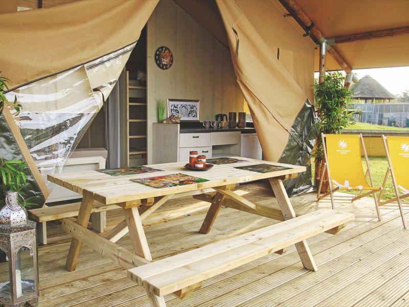 Gemütlicher Zeltplatz mit Holztisch, Bänken und gelben Liegestühlen auf Holzboden.