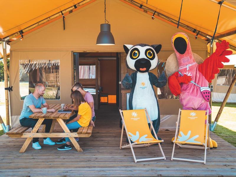 Kinder sitzen an einem Holztisch vor einem Zelt, daneben zwei Personen in Tierkostümen neben Liegestühlen.