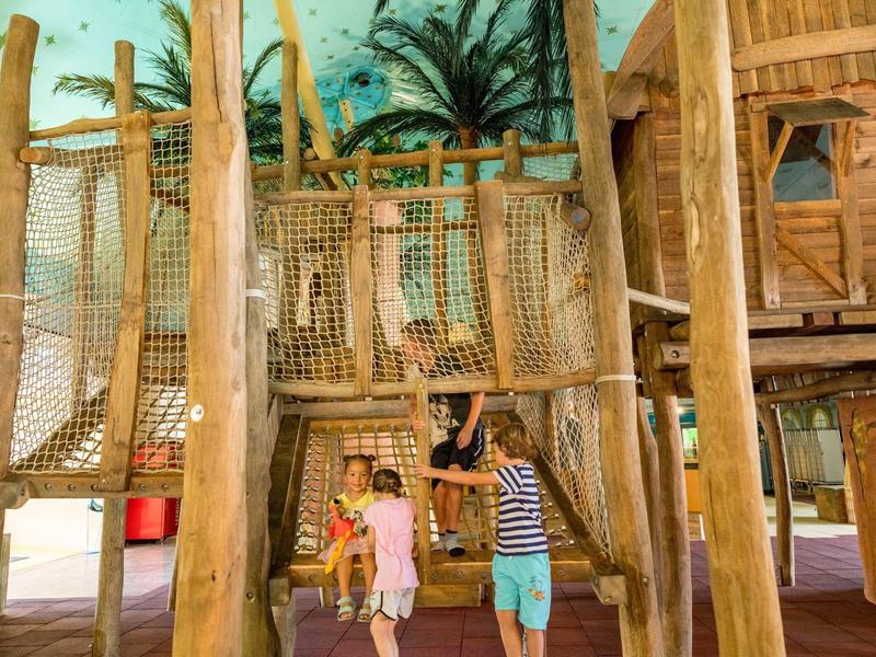 Kinder spielen in einem hölzernen Indoor-Spielplatz mit Kletternetzen und Baumhaus.