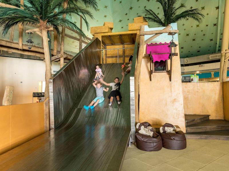 Kinder rutschen eine große Rutsche in einem Innen-Spielbereich mit Palmen und Kissen.
