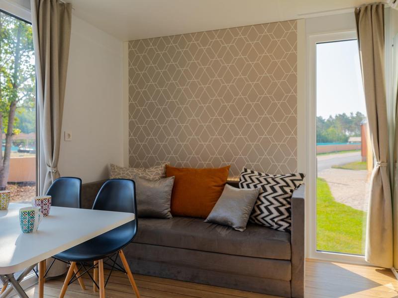 Gemütliches Wohnzimmer mit Grau Sofa, Kissen, Tisch und Stühlen, viel Tageslicht.