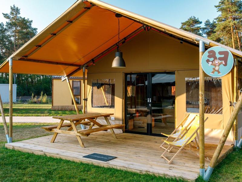 Modernes Safari-Zelt mit überdachter Veranda, Holztisch und Liegestühlen auf einer Wiese.