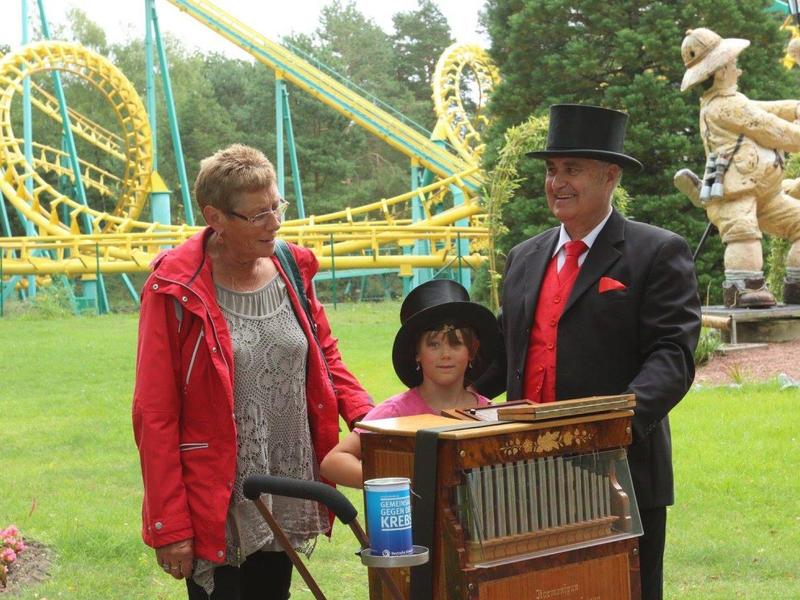 Drei Personen in Freizeitpark, zwei Erwachsene mit Zylinder, Kind mit Hut vor Drehorgel, Achterbahn im Hintergrund.