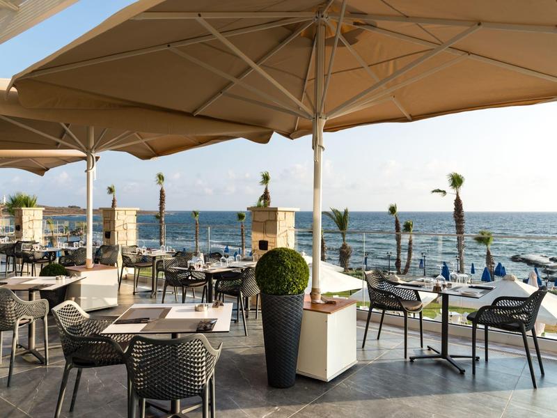 Terrasse mit Tischen, Stühlen und großen Sonnenschirmen am Meer bei klarem Wetter.