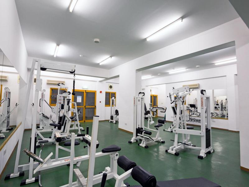 Fitnessraum mit weißen Wänden, grünen Boden, mehreren Trainingsgeräten und Spiegeln an der Wand.