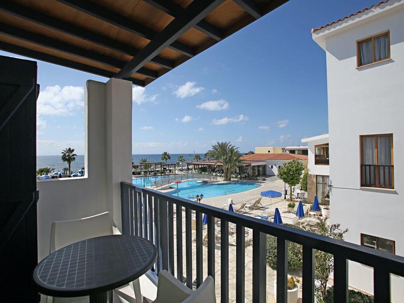 Blick von Balkon mit Tisch und Stuhl auf Pool, Palmen und weiße Gebäude bei blauem Himmel.